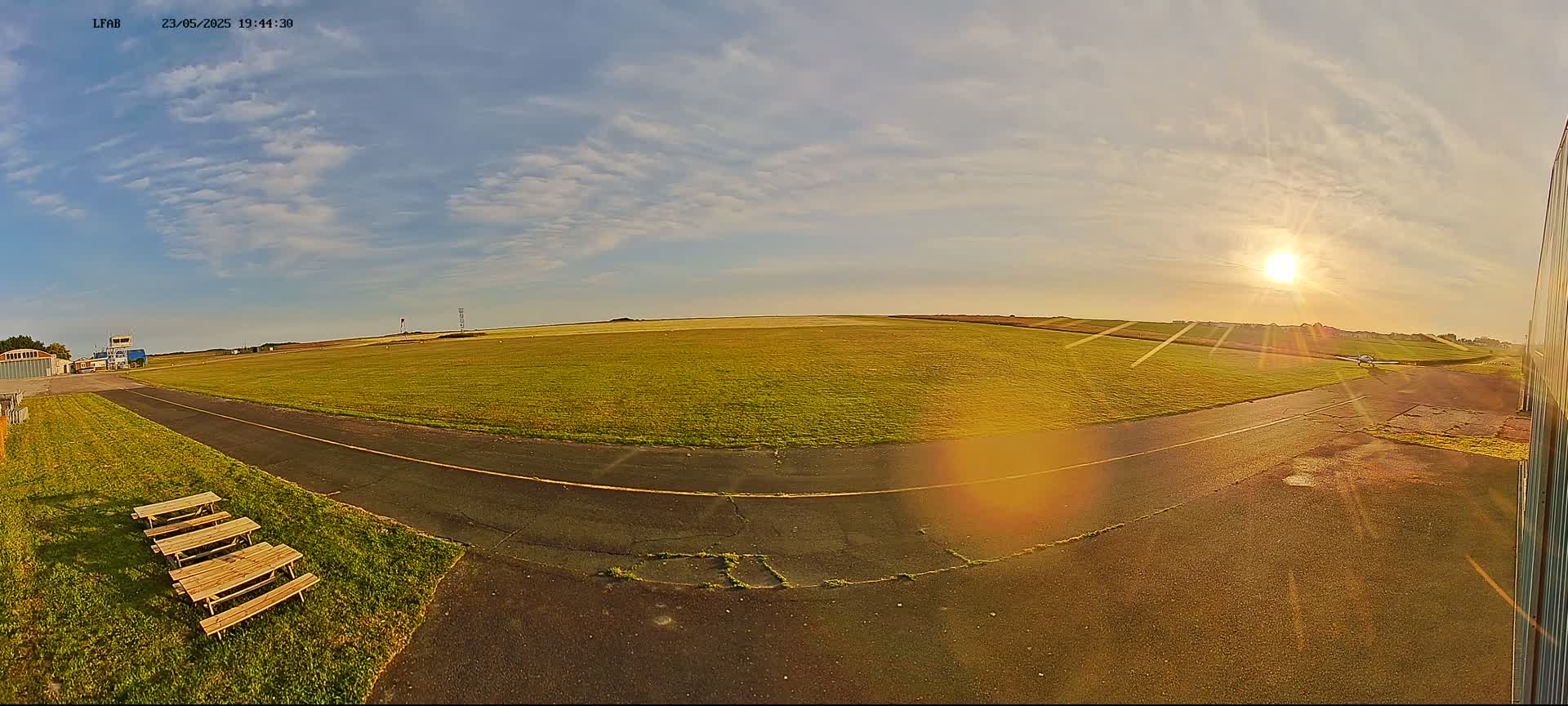 A grassy airfield with a paved runway and picnic tables is bathed in the golden light of a setting sun under a partly cloudy sky.