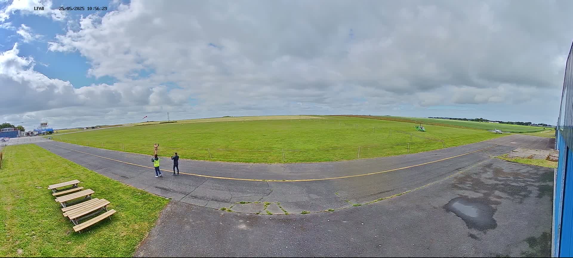 Three people stand near a paved area of an airfield on a partly cloudy day, with a small aircraft visible in the distance and a large grassy field stretching beyond.