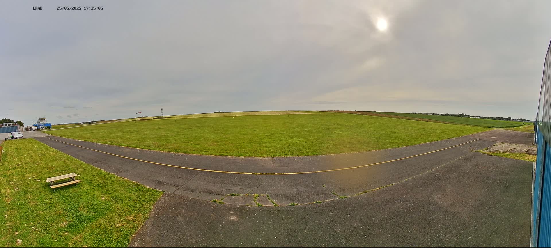 A grassy airfield with a paved taxiway is visible under an overcast sky with the sun barely visible.