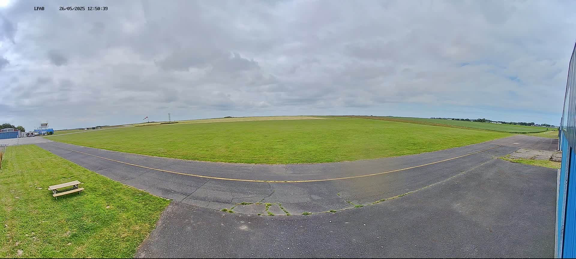 A grassy airfield with a paved taxiway curves toward a small building under a mostly cloudy sky.