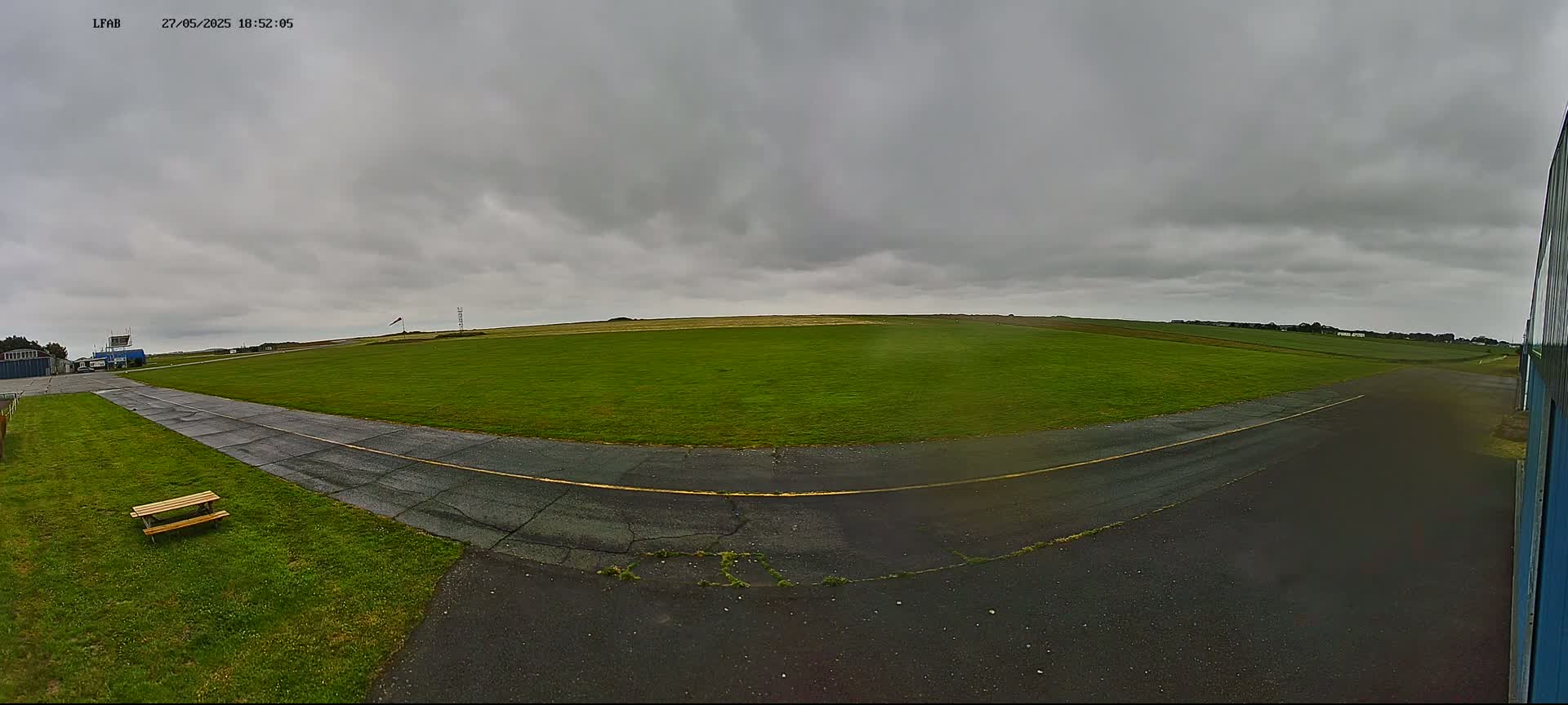 A grassy airfield under an overcast sky shows a paved taxiway curving toward a small building and a picnic bench.