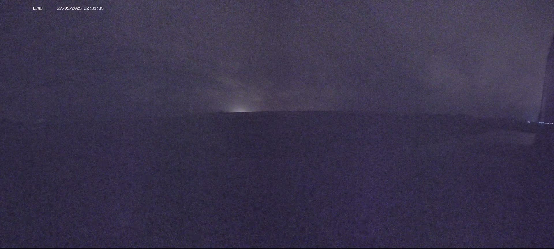 A dark, nighttime outdoor scene shows a flat, low horizon under a cloudy, possibly stormy sky with a faint light source visible in the distance.