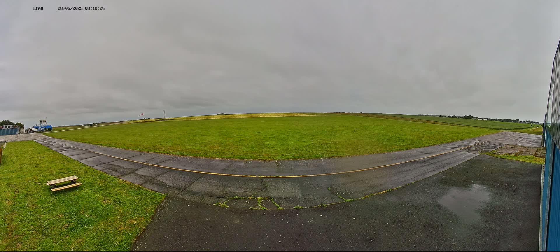 A grassy airfield with a paved runway and a picnic table is visible under an overcast sky.