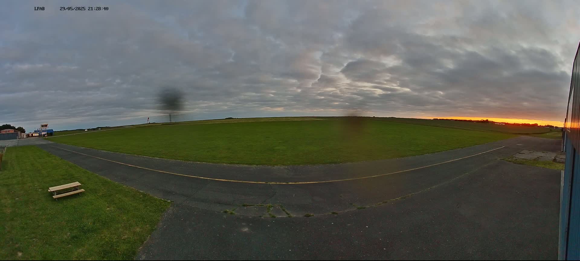 A grassy airfield with a paved taxiway is visible at sunset under a cloudy sky.