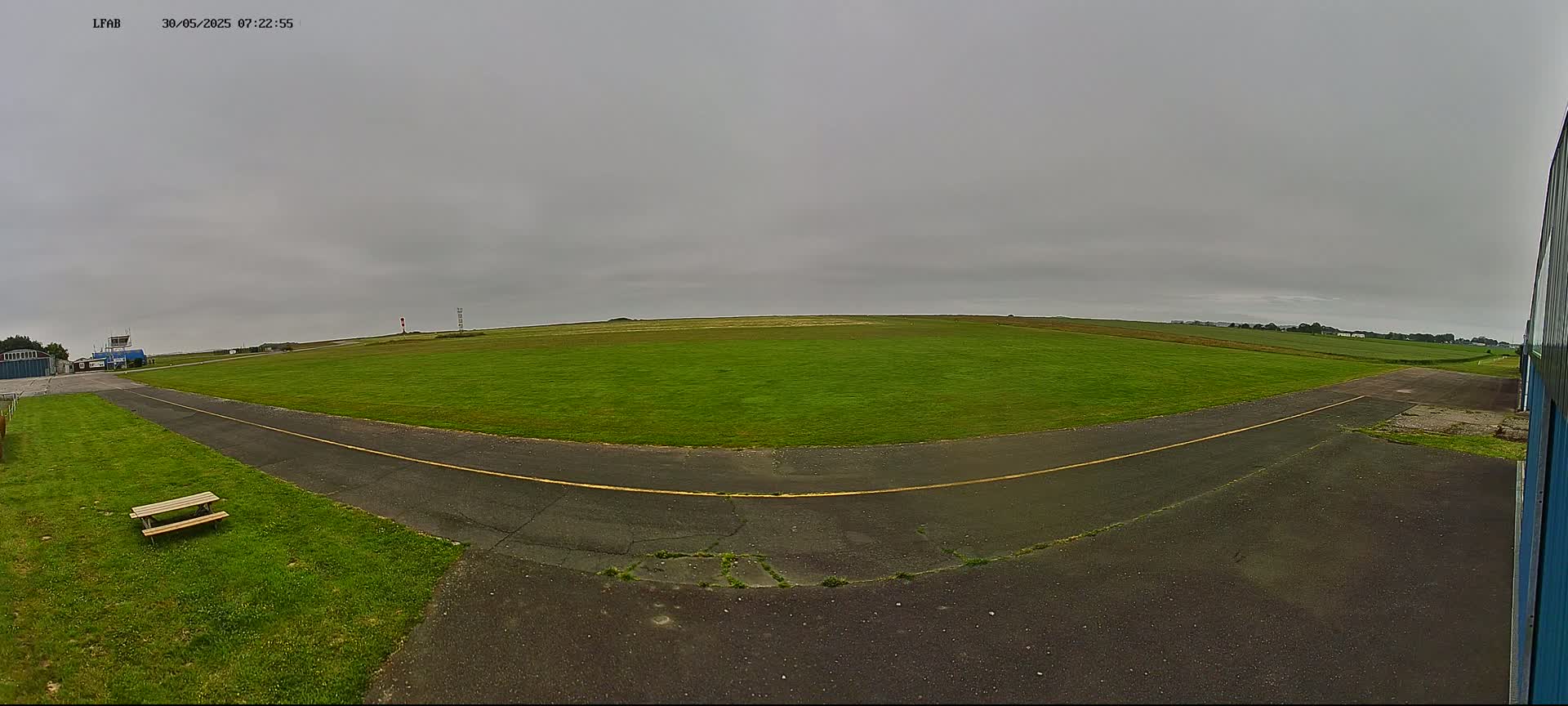A grassy airfield with a paved taxiway under an overcast sky is seen from a low vantage point near a hangar.