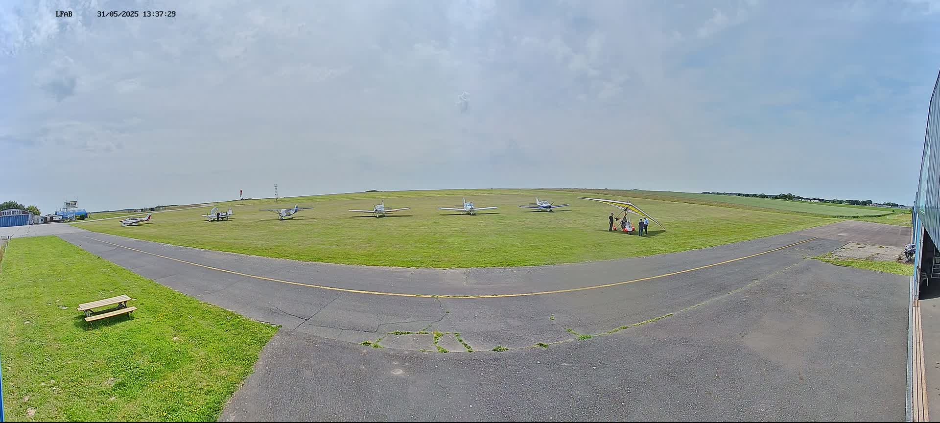 Several small airplanes and a hang glider are parked on a grassy airfield under a partly cloudy sky.