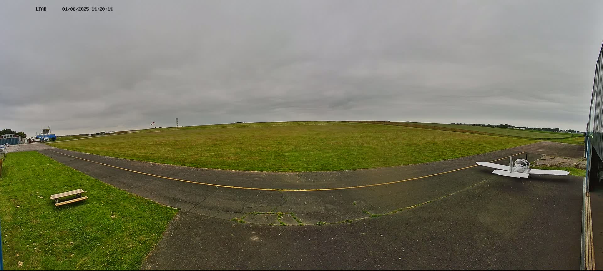 A small, white airplane sits on a tarmac next to a grassy airfield under an overcast sky.
