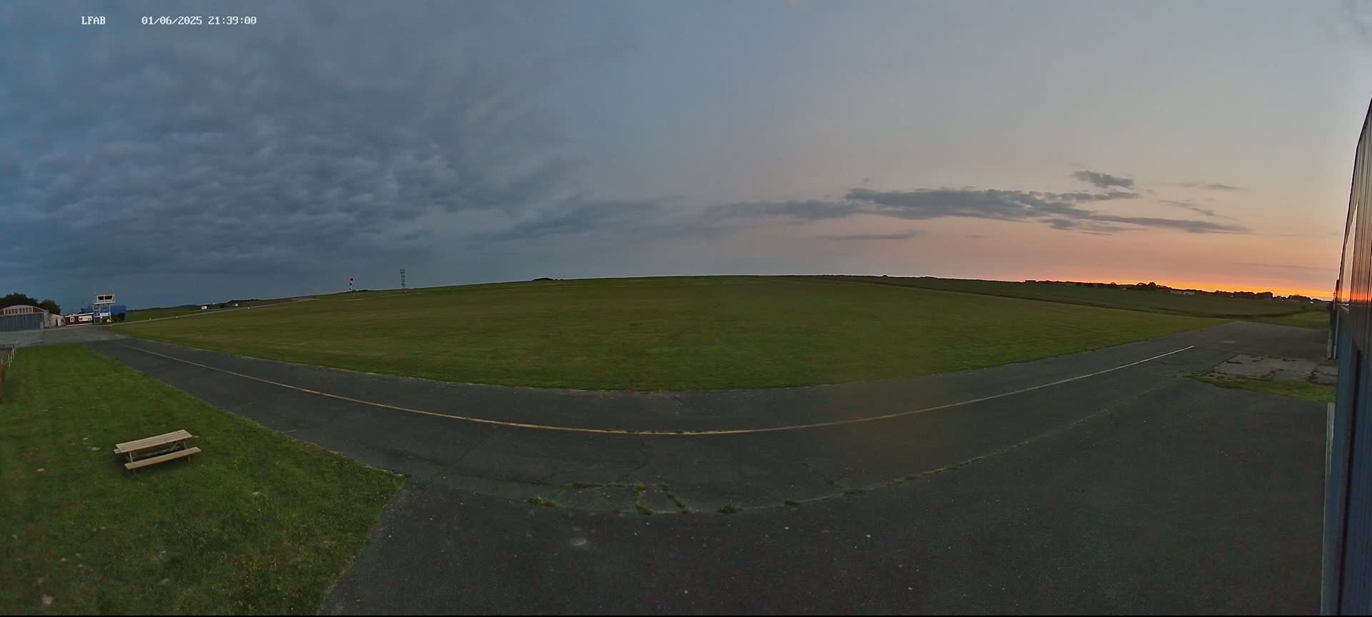 A wide, grassy airfield at sunset under a partly cloudy sky, featuring a paved taxiway and a picnic table.