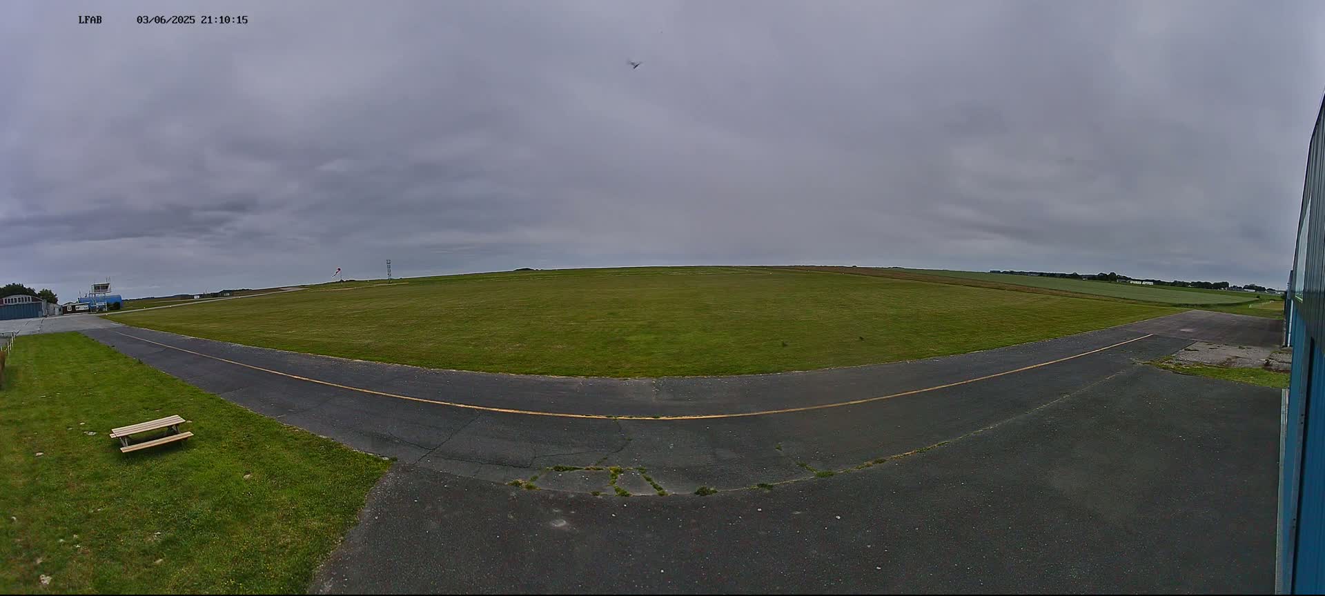 A small airfield with a grassy field, a paved taxiway, and a single picnic table is visible under an overcast sky.