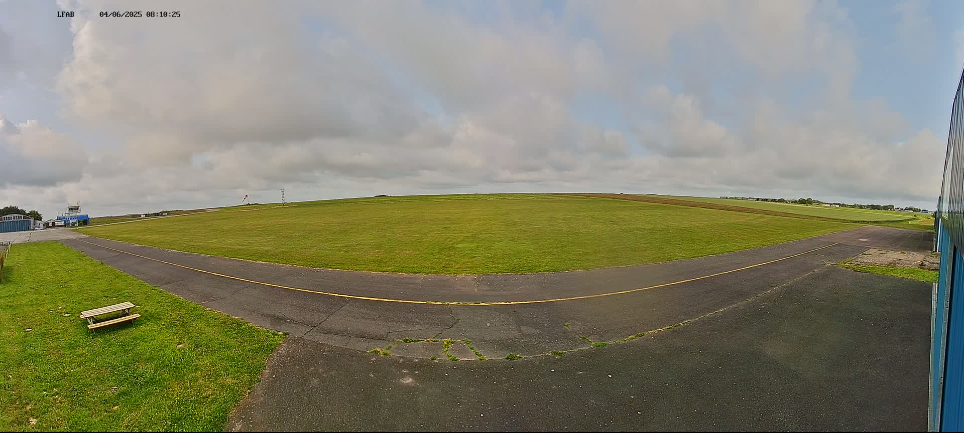 A grassy airfield with a paved taxiway and a small picnic table is visible under a mostly cloudy sky.
