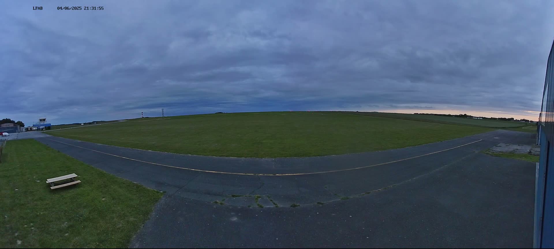 A grassy airfield with a paved taxiway is visible under a partly cloudy, dusky sky.