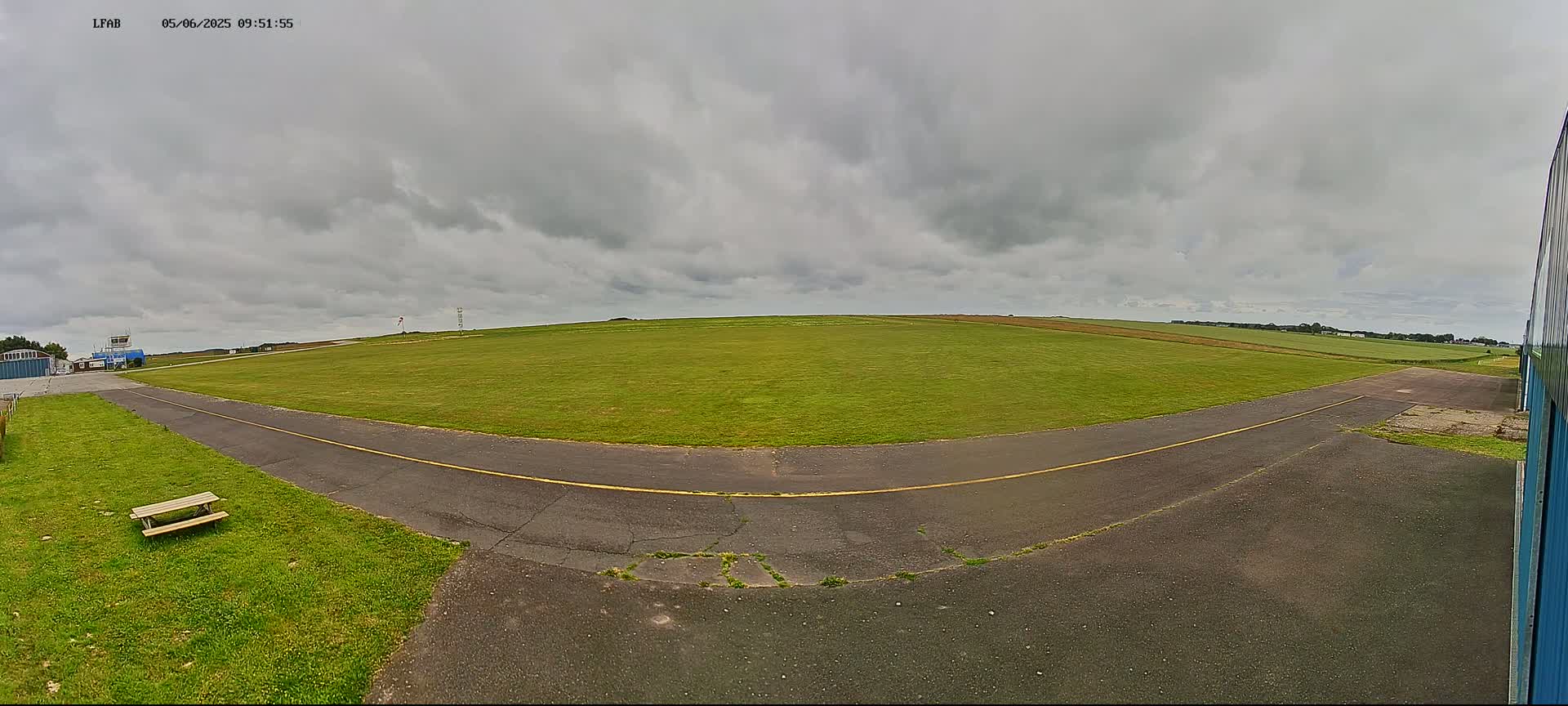 A grassy airfield under a cloudy sky shows a paved taxiway curving toward a hangar.