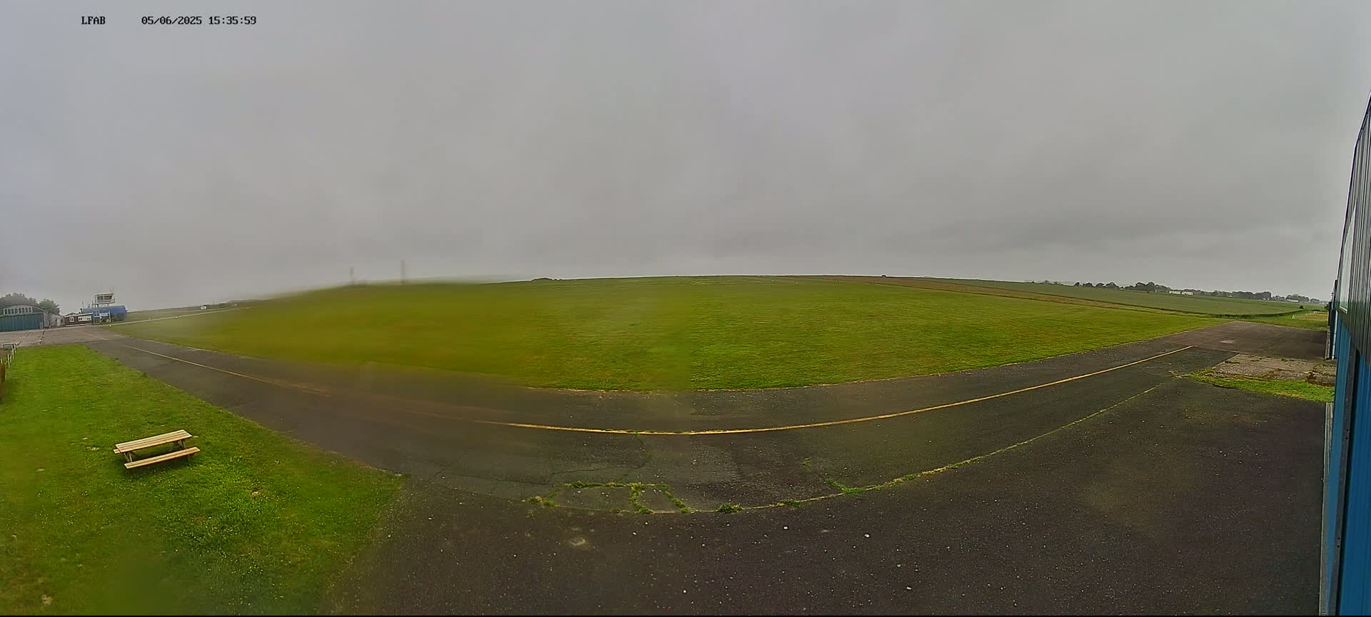 A grassy airfield under an overcast sky is seen from a ground-level perspective, with a paved taxiway leading to a distant horizon.