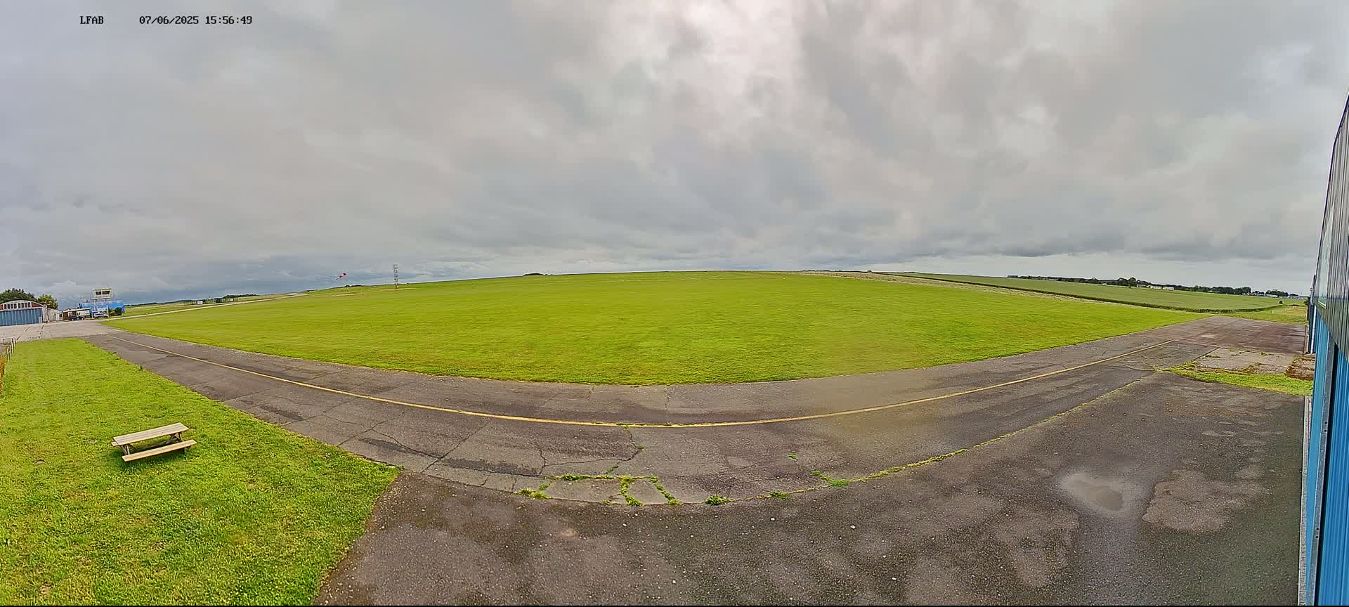 A grassy airfield under an overcast sky shows a paved taxiway curving toward a large, open field.