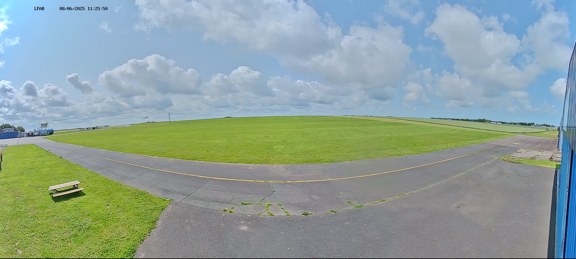 A grassy airfield under a partly cloudy sky shows a paved taxiway curving toward a large green field.