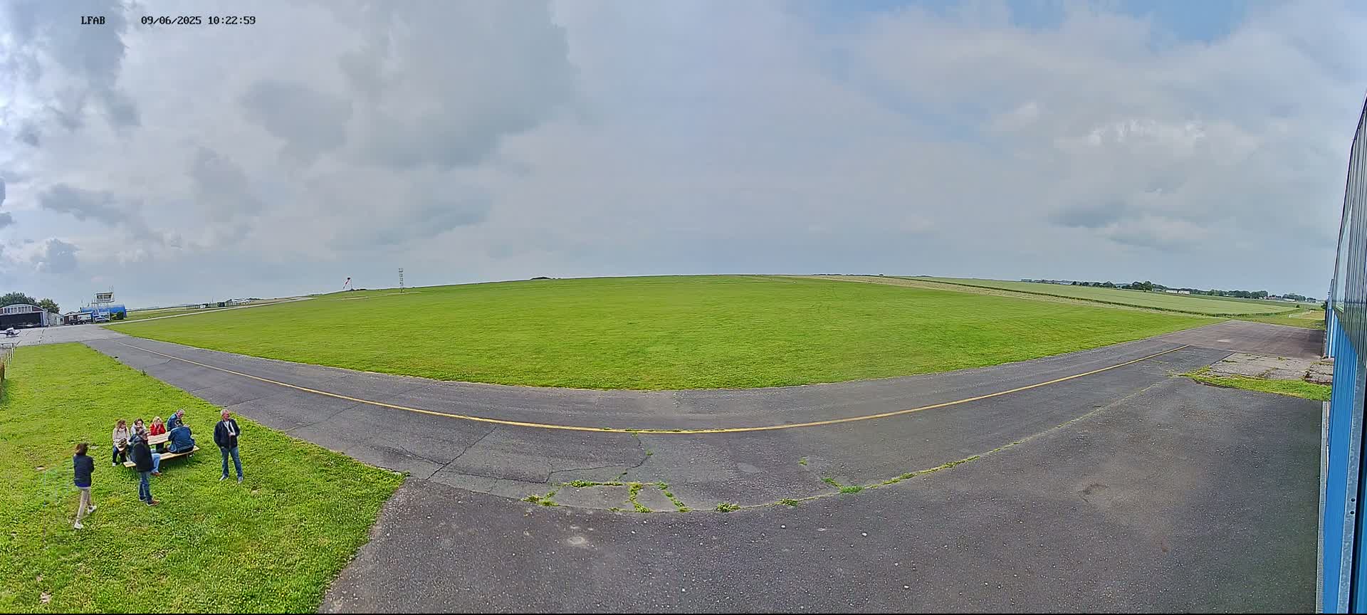A wide, grassy airfield with a paved taxiway curves toward a group of people gathered near a hangar under a partly cloudy sky.