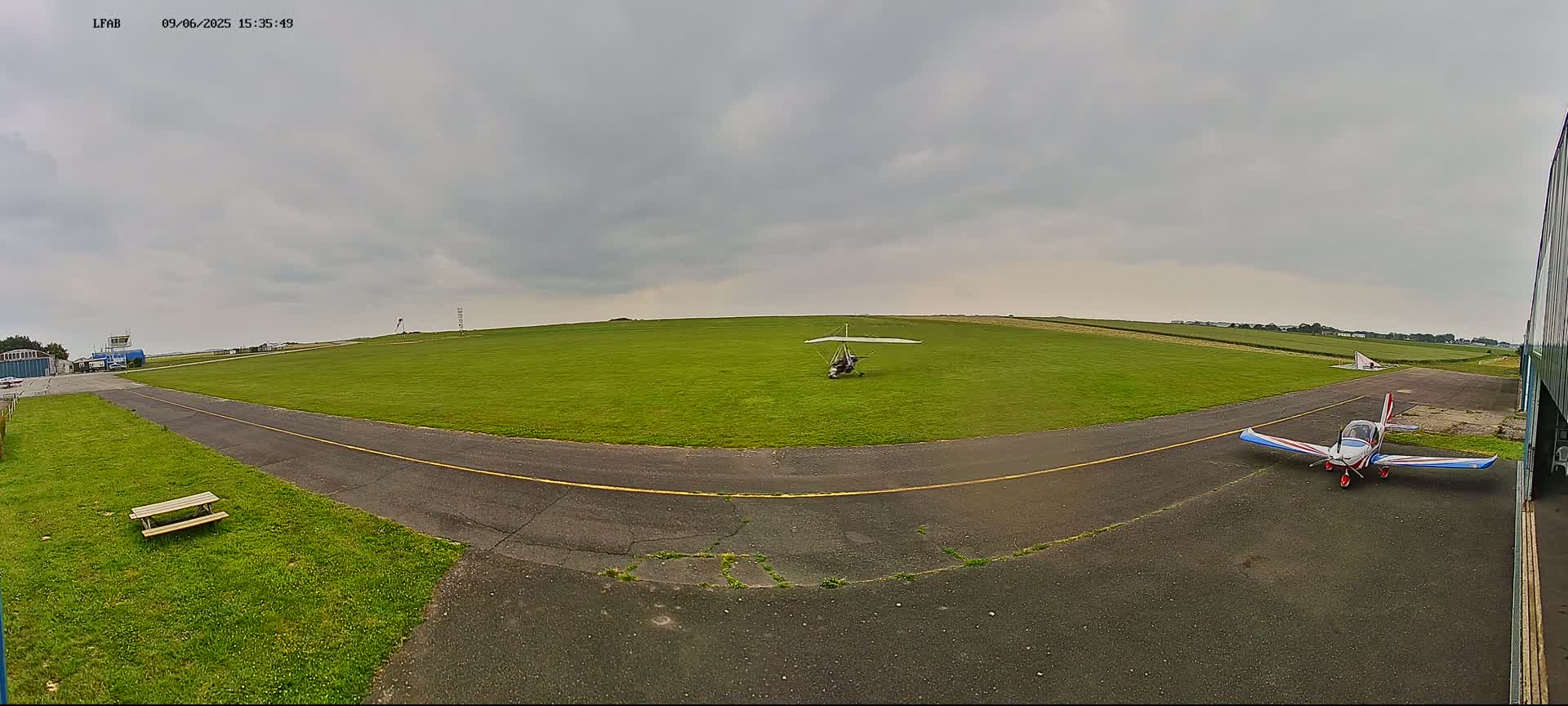 A small airfield with two light aircraft, one on the grass and one on the tarmac, under an overcast sky.