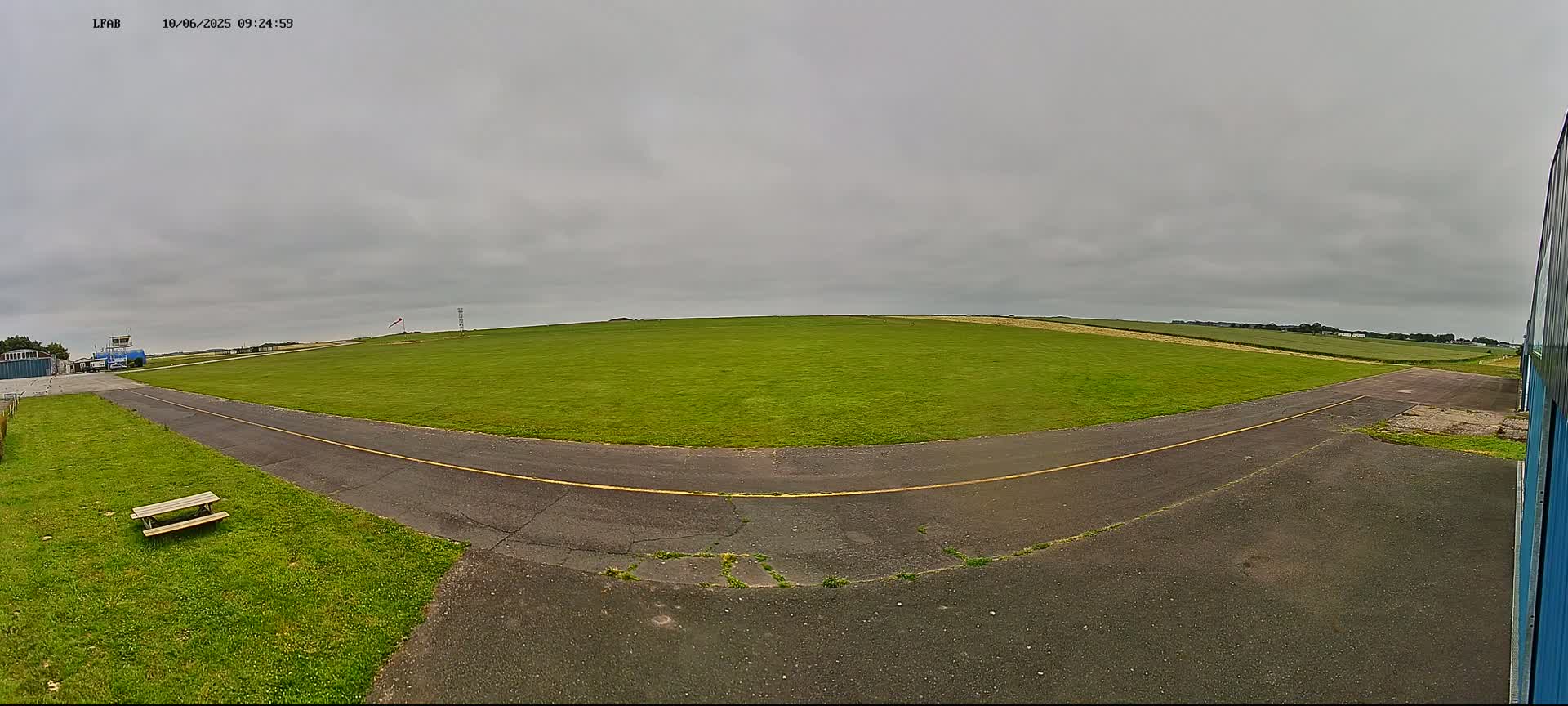 A grassy airfield with a paved taxiway under an overcast sky is visible, featuring a picnic table and a building at the edge.