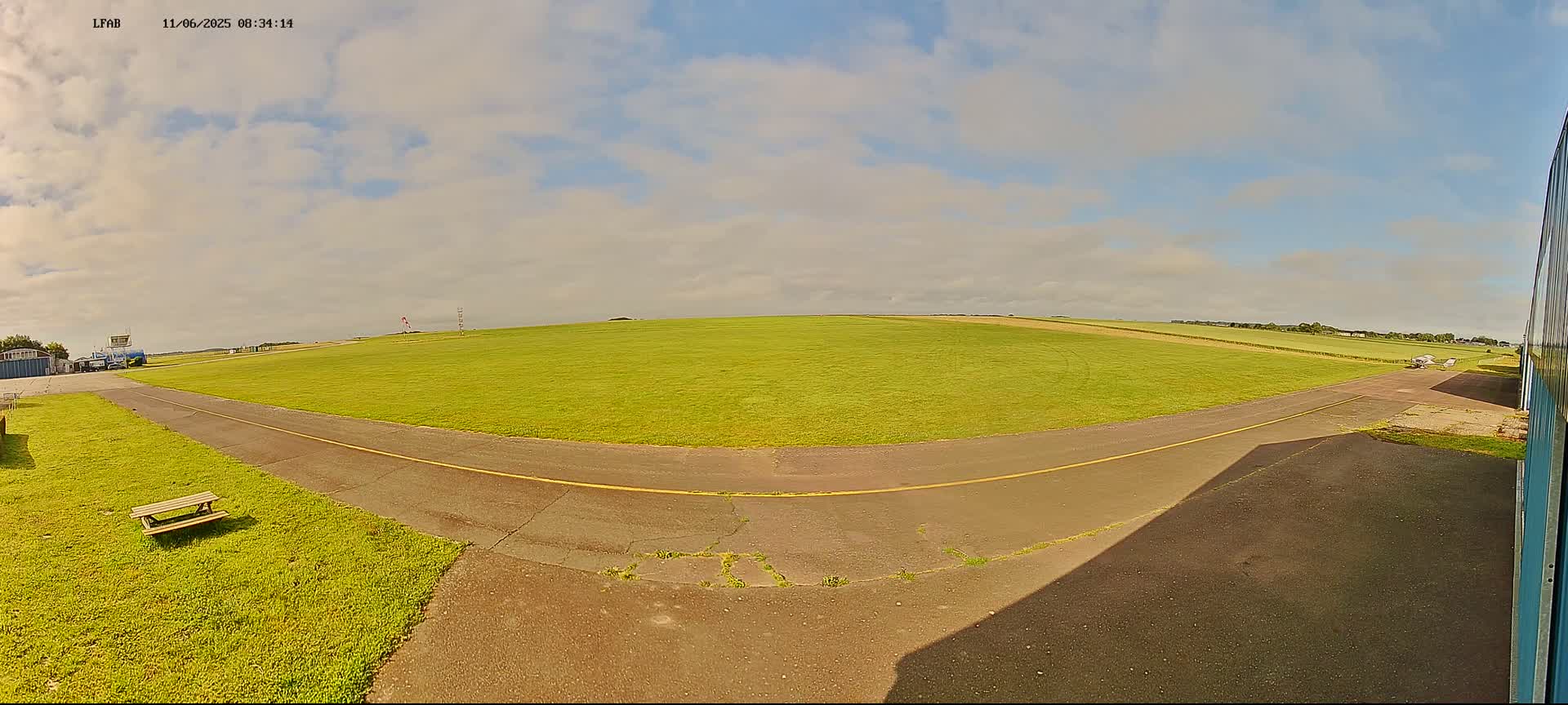 A mostly sunny day at a small airfield shows a grassy runway with a paved taxiway leading to a hangar, and a picnic table sits nearby.