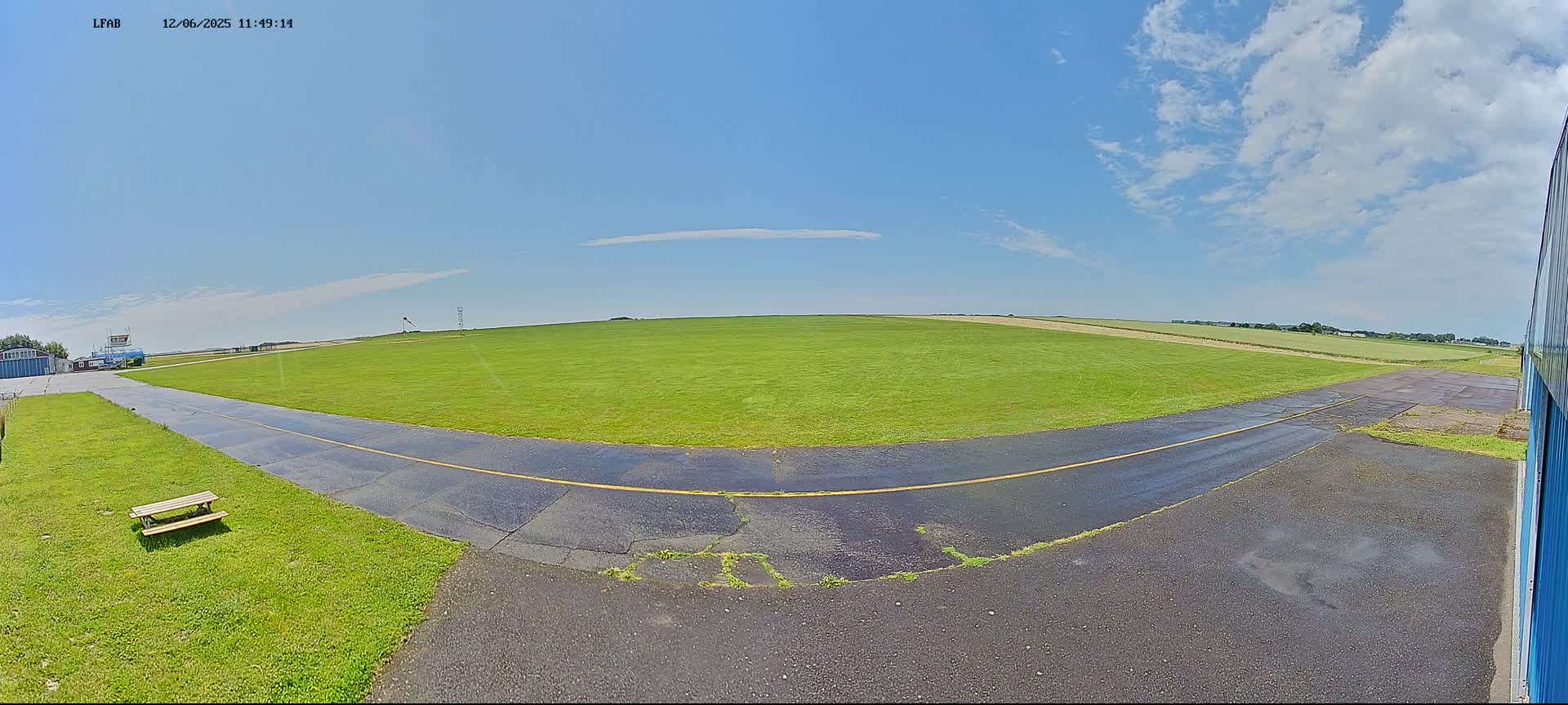 A mostly sunny day reveals a grassy airfield with a paved taxiway curving to the right, a picnic table sits in the grass, and a section of a blue building is visible in the bottom right corner.