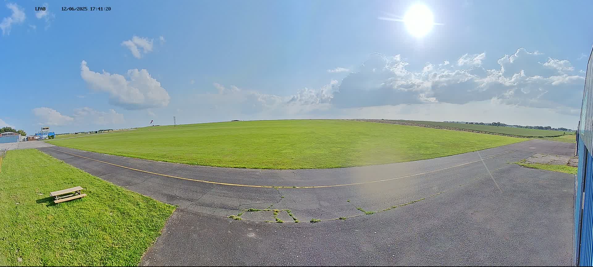 A sunny day with partly cloudy skies shows a grassy airfield with a paved taxiway curving toward a distant horizon.