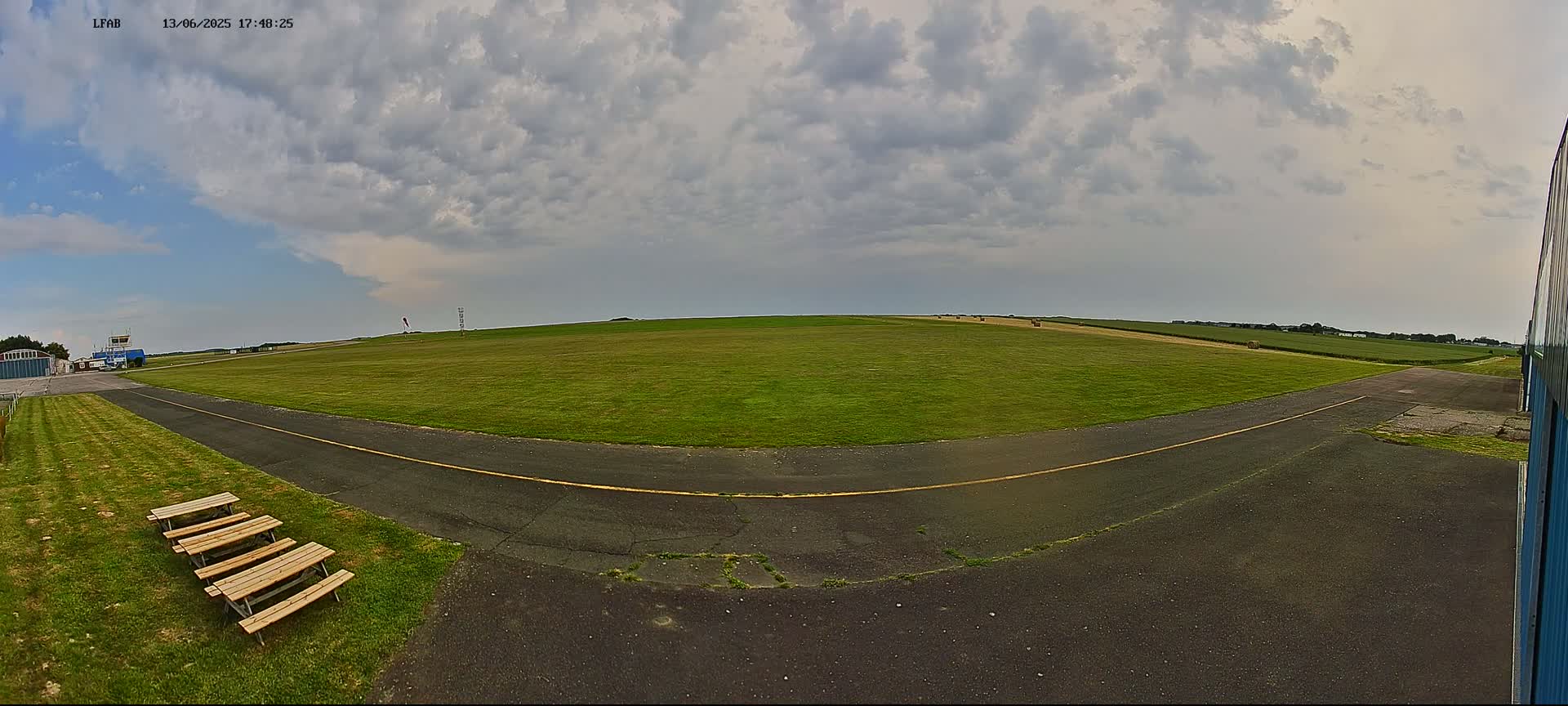 A grassy airfield with a paved taxiway, several picnic tables, and a cloudy sky is visible.
