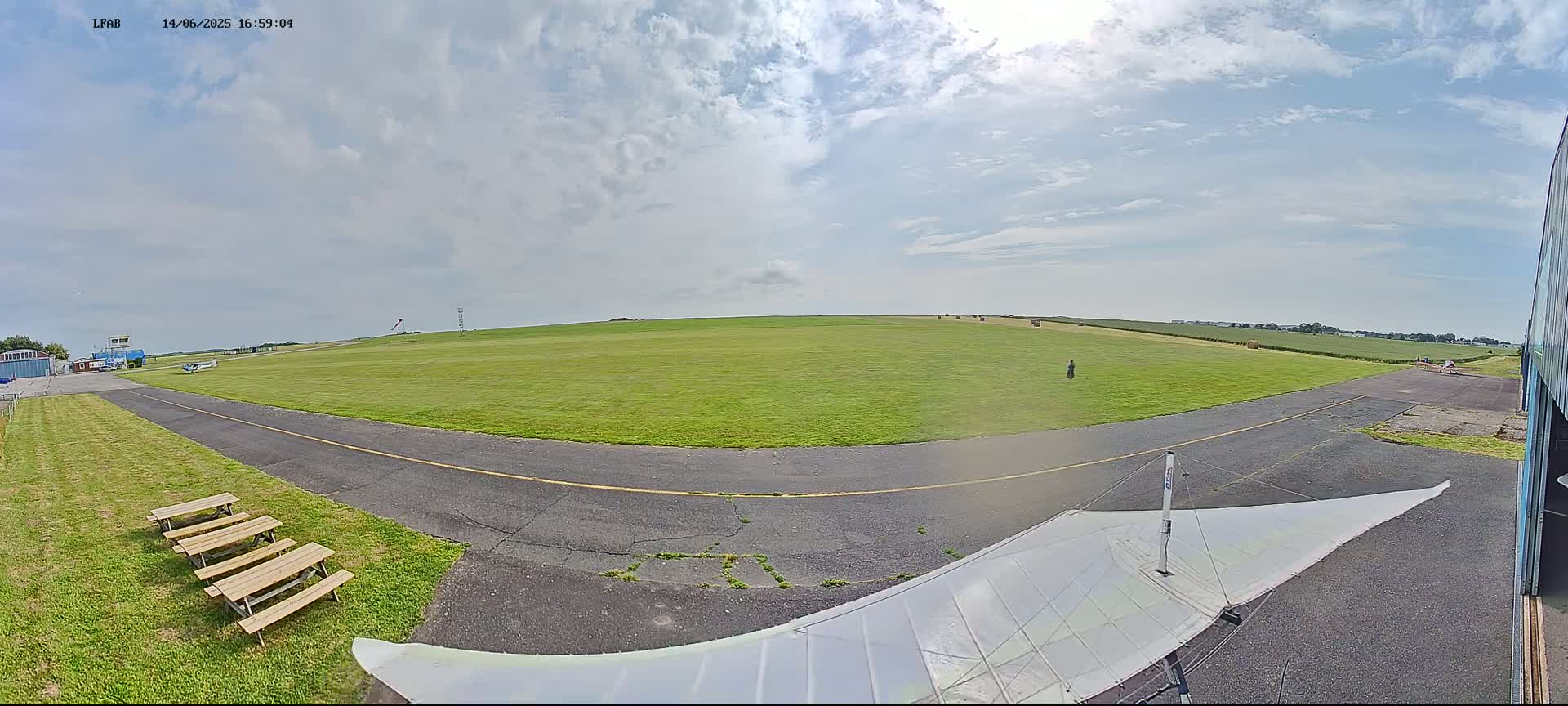 A grassy airfield under a partly cloudy sky is viewed from a low angle, showing a paved taxiway, picnic tables, and a hang glider in the foreground.
