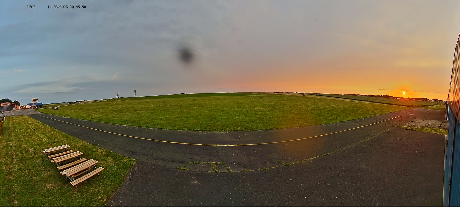 A grassy airfield under a partly cloudy sky is viewed from a low angle, showing a paved taxiway, picnic tables, and a hang glider in the foreground.
