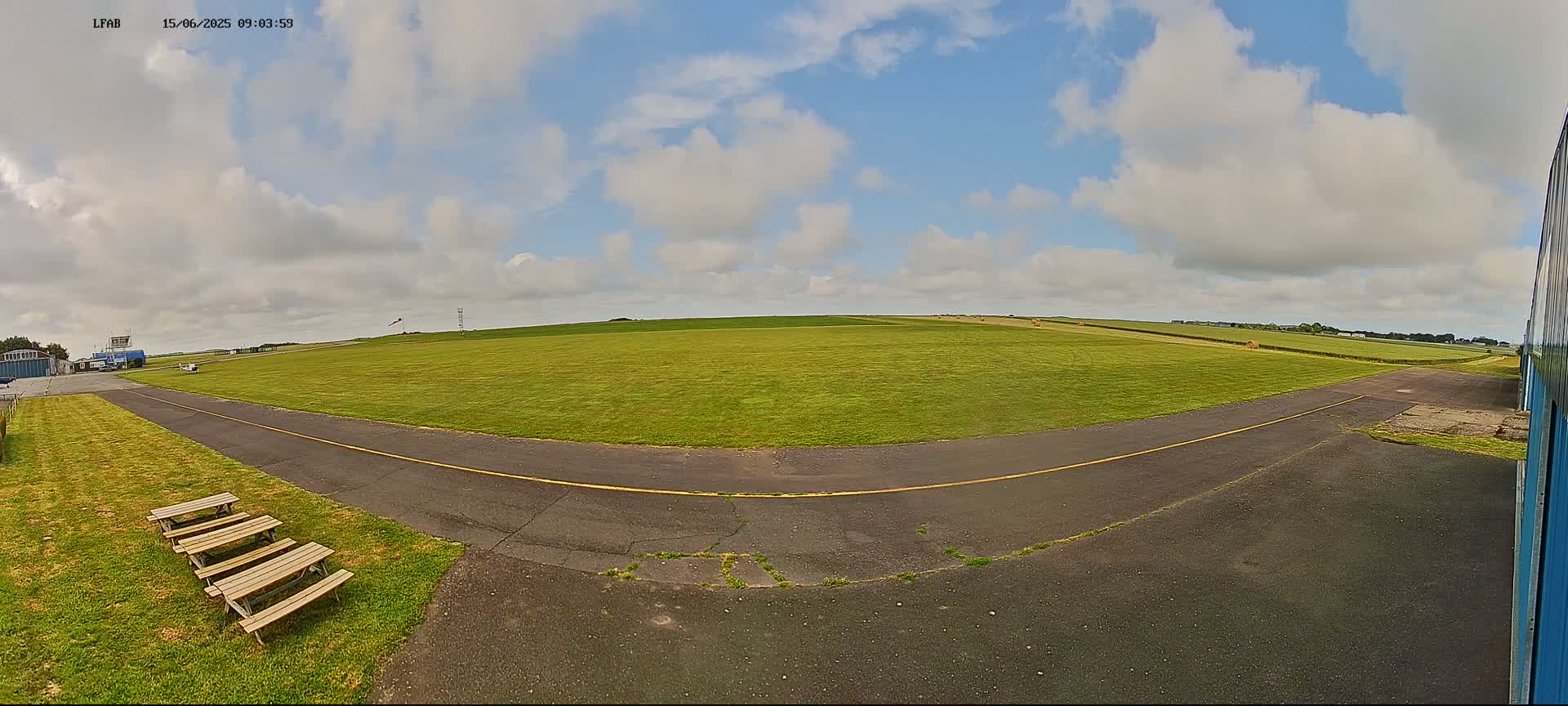 A grassy airfield under a partly cloudy sky features a paved taxiway, several picnic tables, and a hangar partially visible in the right corner.