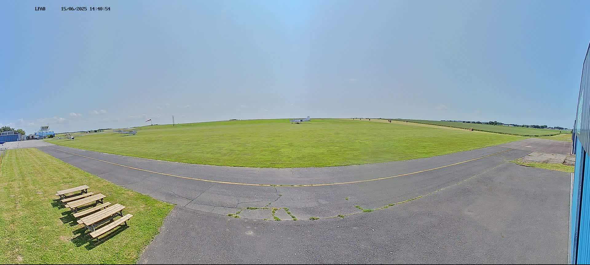 A small airfield on a sunny day with several small airplanes on a grassy field, a paved taxiway, and picnic tables near a hangar.
