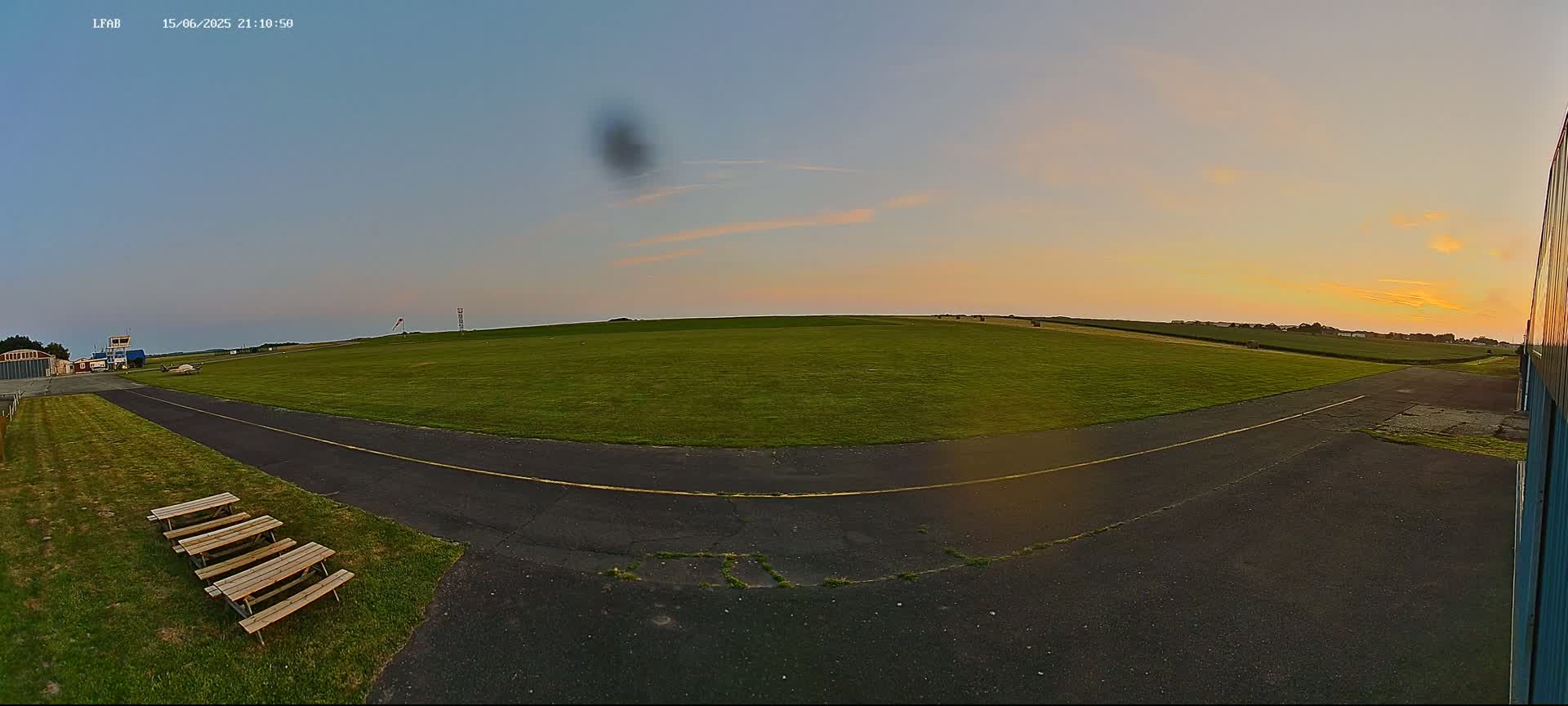 A small airfield on a sunny day with several small airplanes on a grassy field, a paved taxiway, and picnic tables near a hangar.