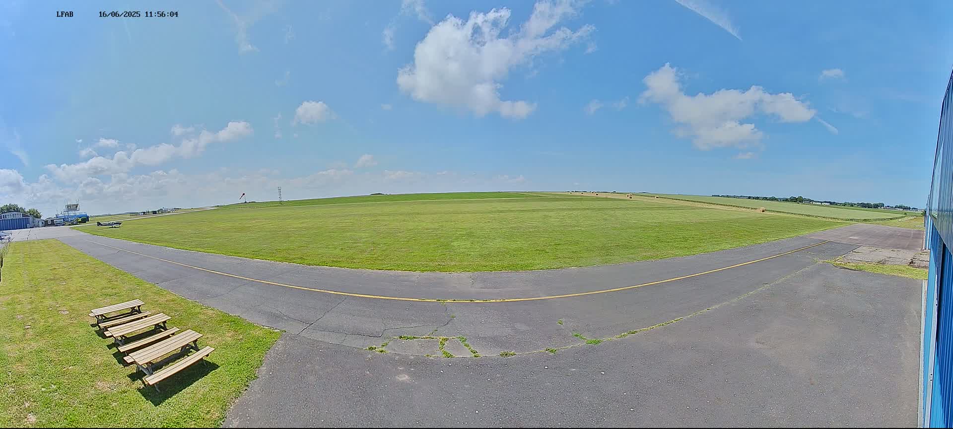 A paved taxiway curves around a grassy airfield under a mostly sunny sky with scattered cumulus clouds.