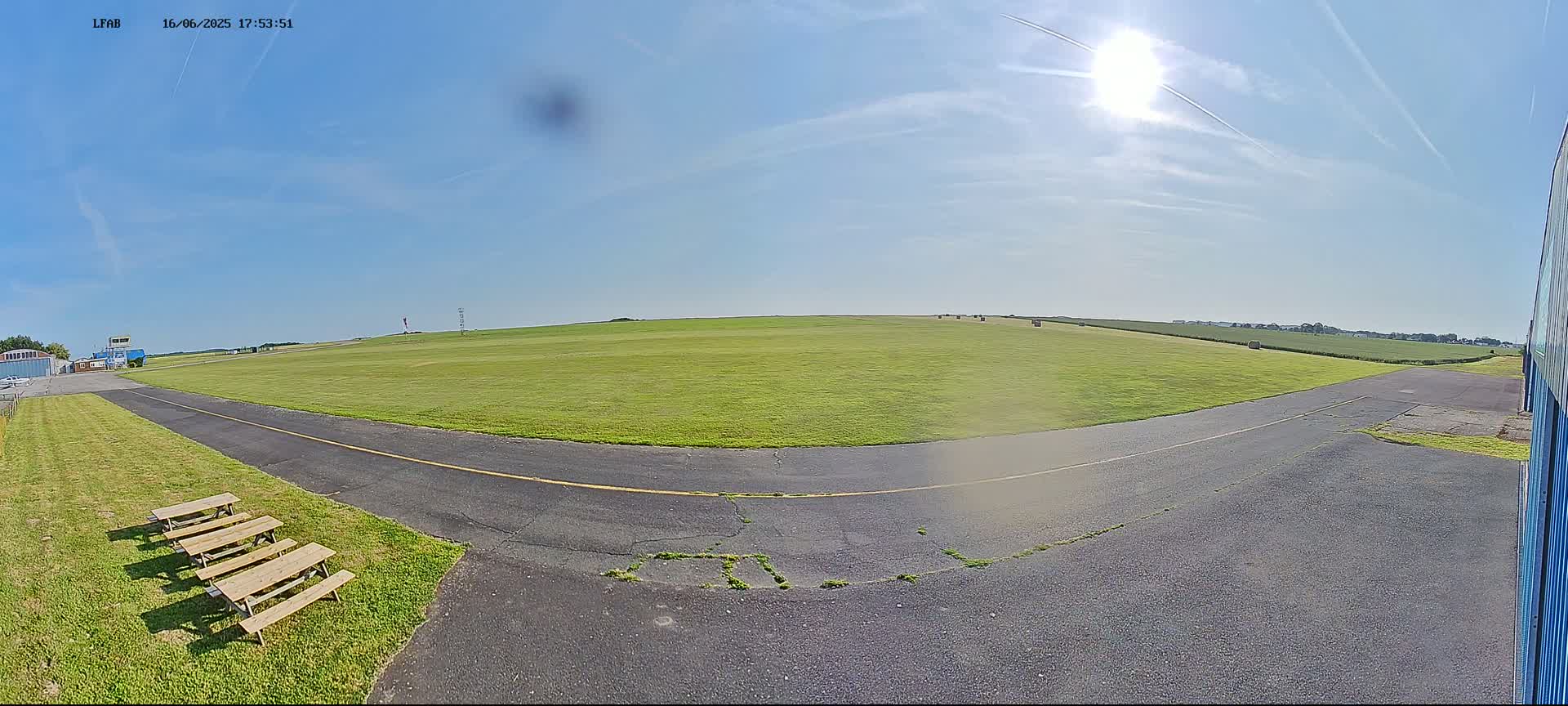 A sunny day at a grassy airfield shows a paved taxiway curving toward a large green field with hay bales in the distance.