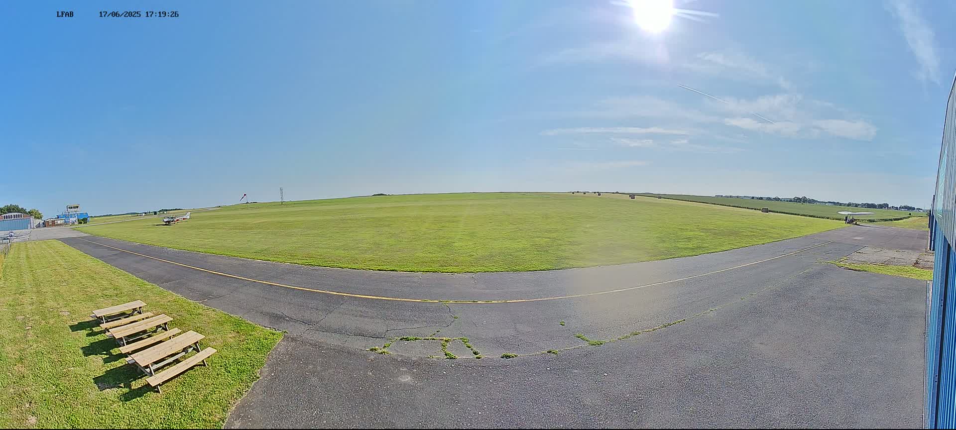 A sunny day at a small airfield shows a grassy field with hay bales in the distance, a paved taxiway, and several picnic tables.