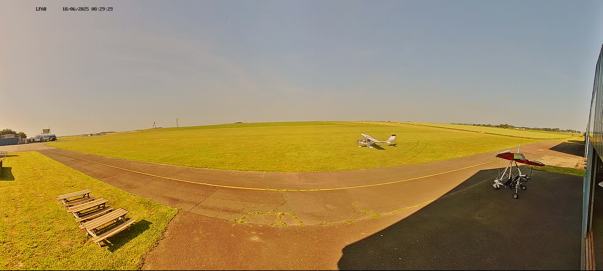 A small airplane and a hang glider sit on a grassy airfield under a clear, sunny sky.