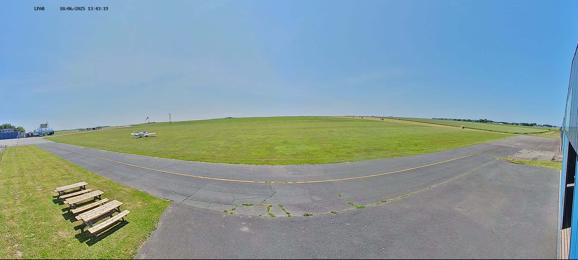 A small airplane sits on a grassy airfield on a sunny day, with several picnic tables near a paved taxiway.