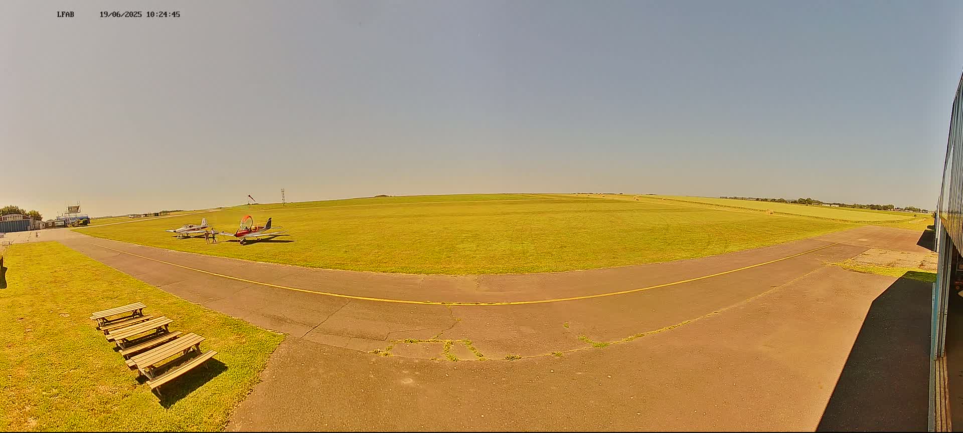 A sunny day at a small airfield shows two small airplanes parked on the grass near a paved taxiway, with picnic tables nearby.