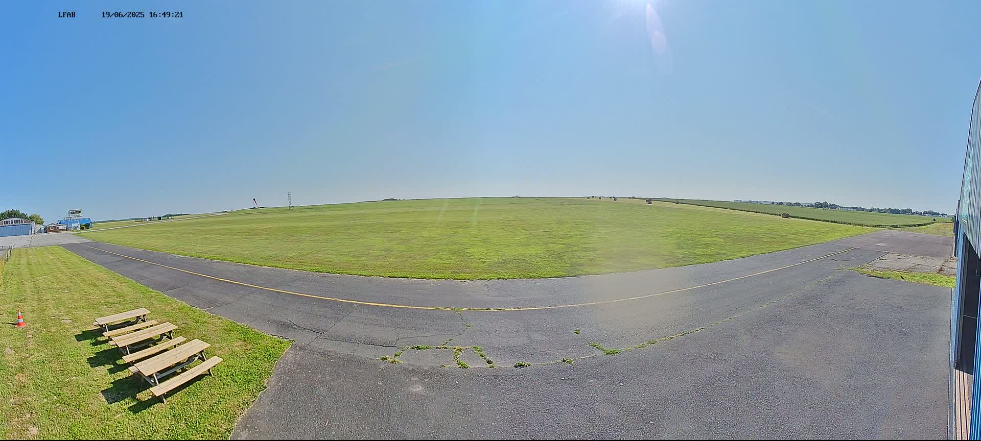 A wide, grassy airfield under a clear, sunny sky is shown, with a paved taxiway curving toward a distant horizon where hay bales are visible in a field.
