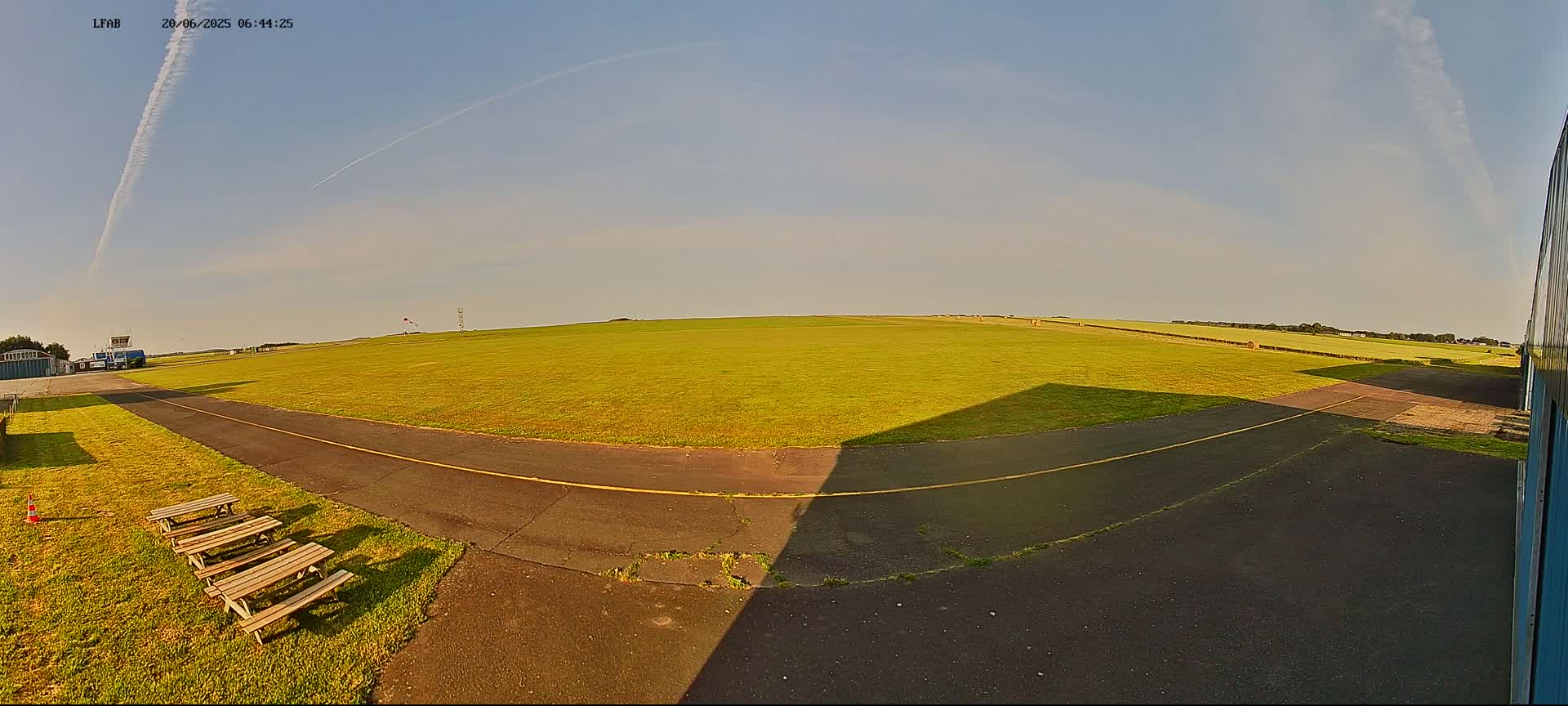 A grassy airfield under a clear, sunny sky shows a paved taxiway with picnic tables near a hangar.