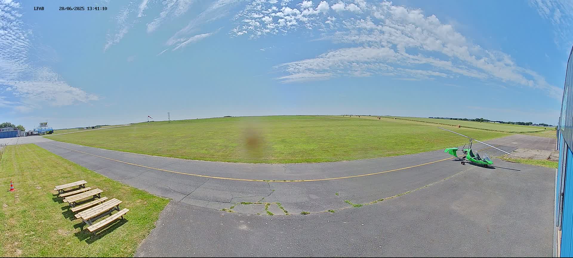 A bright, sunny day at a small airfield shows a green gyrocopter parked on the tarmac near several picnic tables, with a grassy airfield stretching into the distance.