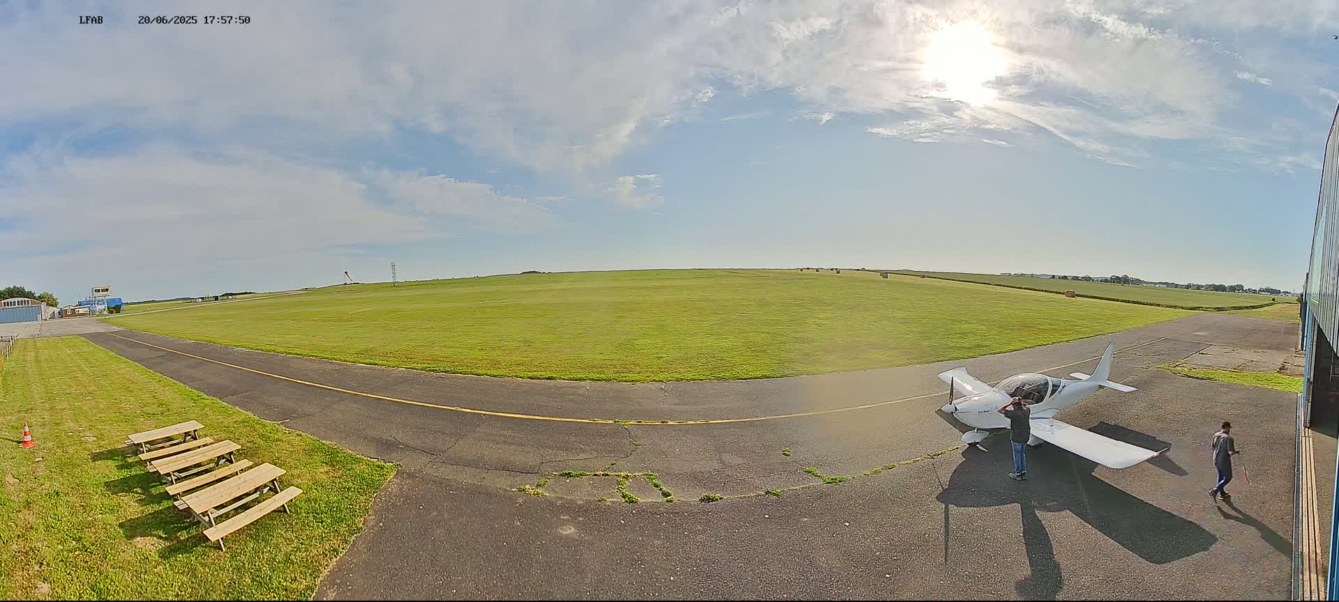 A small white airplane sits on a grassy airfield under a mostly sunny sky with scattered clouds, near two people and several picnic tables.