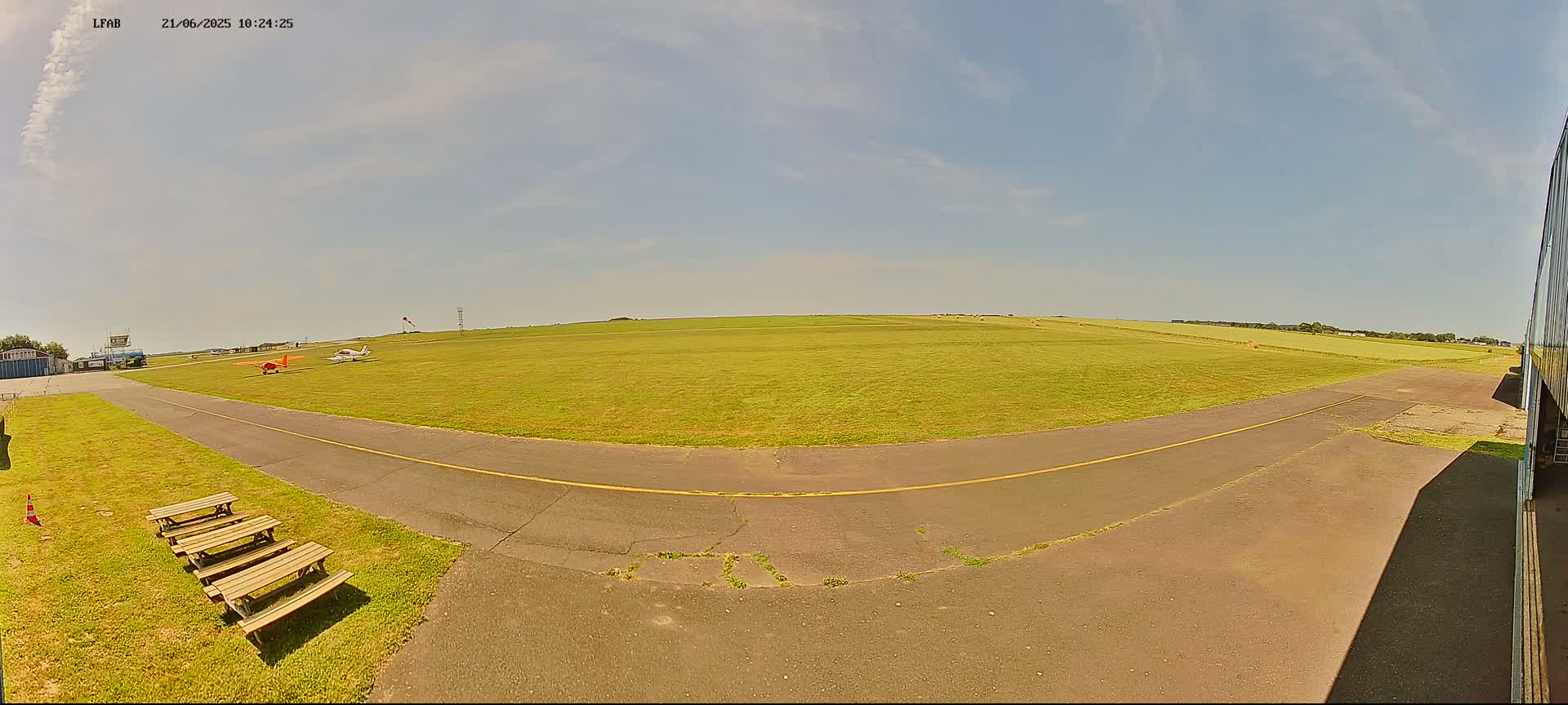 Two small airplanes sit on a grassy airfield under a mostly sunny sky, near a paved taxiway and picnic tables.