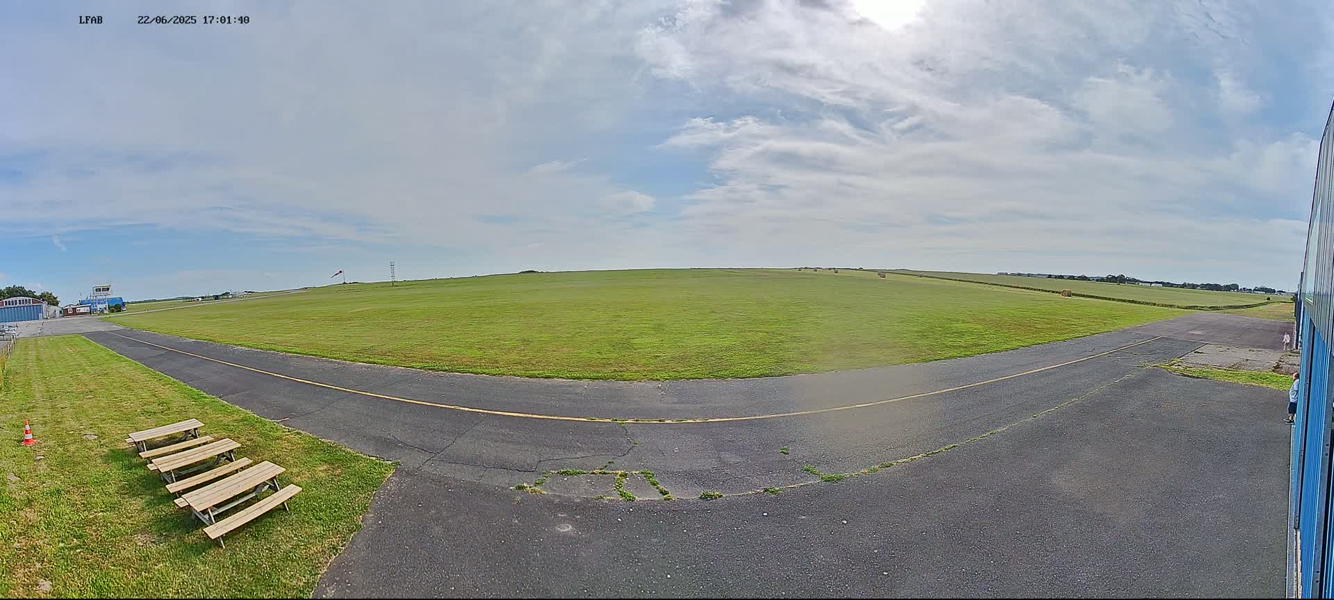 A mostly sunny day reveals a grassy airfield with a paved taxiway, several picnic tables, and a distant building.