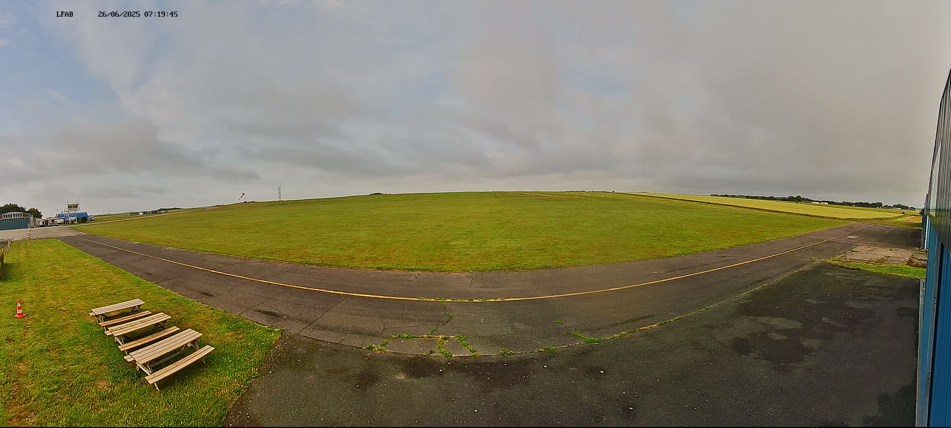 A grassy airfield with a paved taxiway curves toward a large green field under an overcast sky.