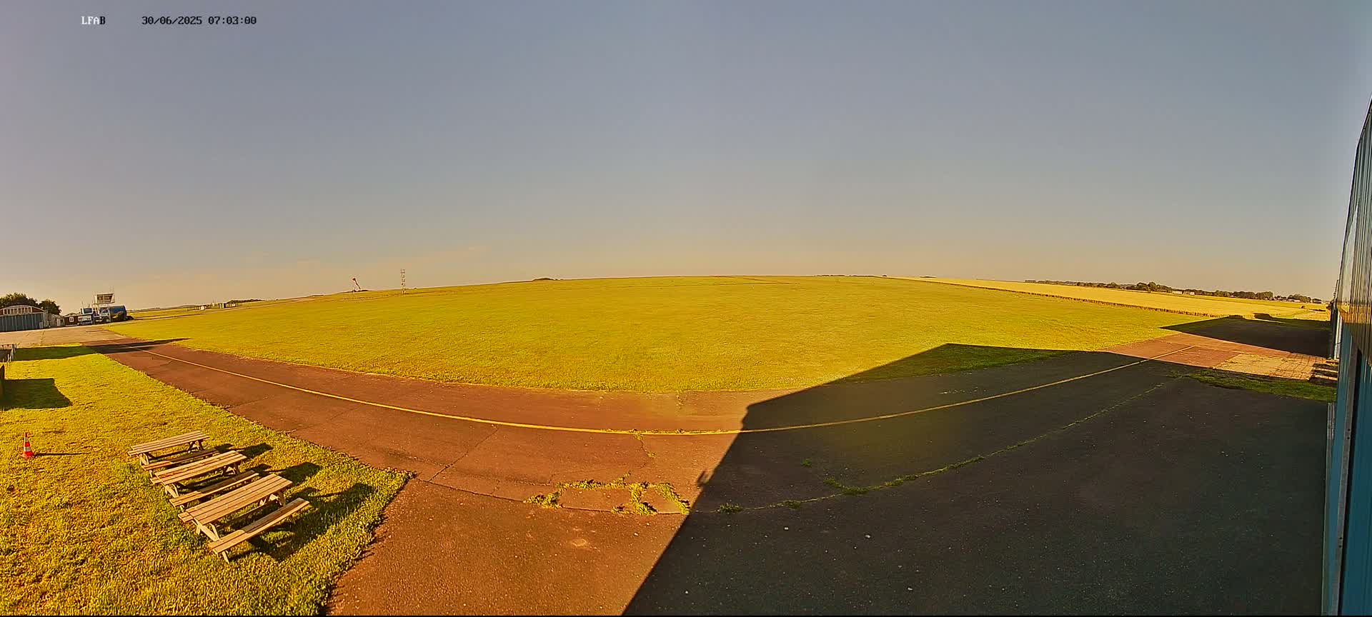 A grassy airfield under a clear, sunny sky features a paved taxiway, picnic tables, and a building casting a long shadow.