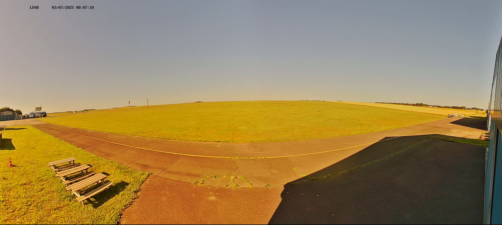 A grassy airfield with a paved taxiway and picnic tables is visible under a clear, sunny sky.