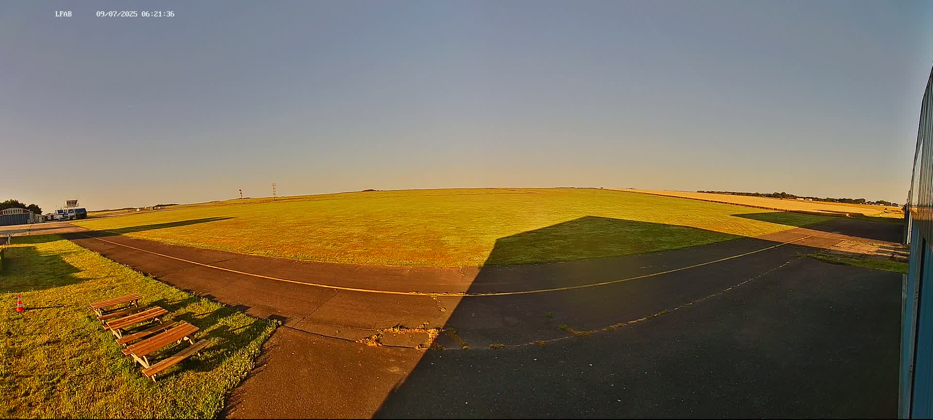 A grassy airfield under a clear, bright sky, with picnic tables and a paved area nearby.