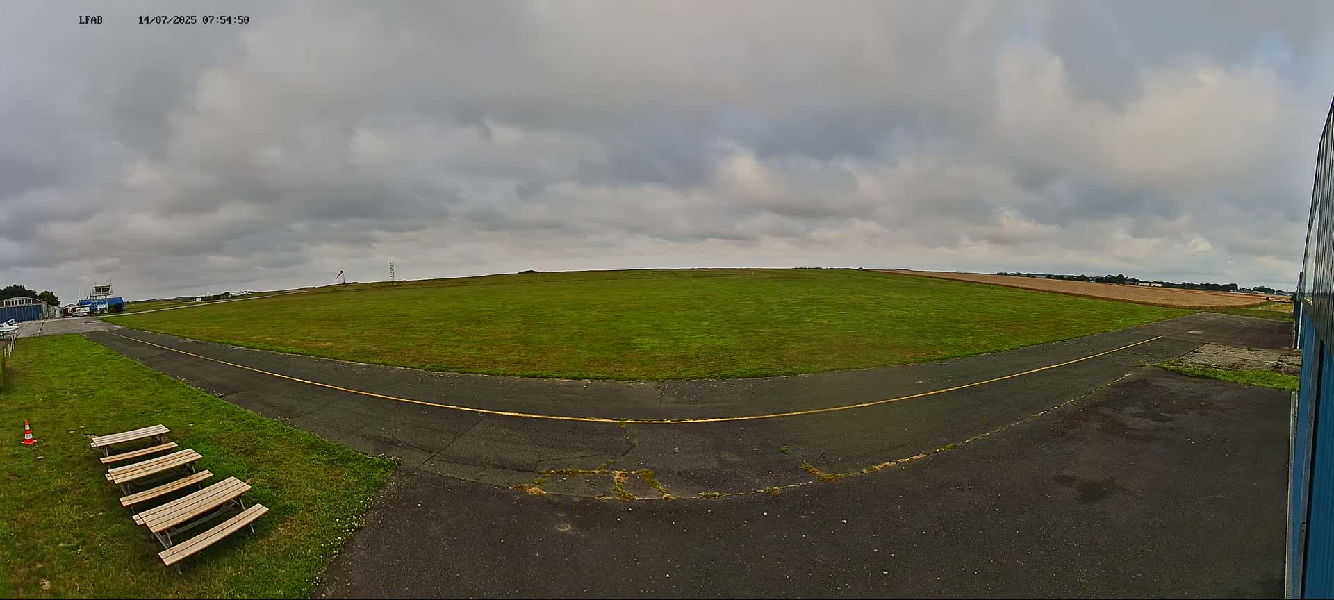 A grassy airfield with a paved taxiway and several picnic tables is visible under a mostly cloudy sky.