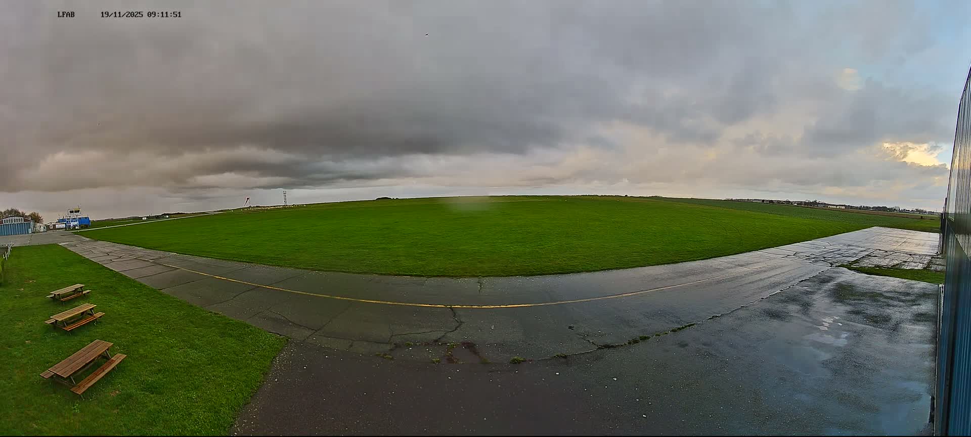 A wide panoramic view of an airfield shows wet tarmac and lush green fields under a heavily overcast and gloomy sky, with some brighter light attempting to break through the clouds on the horizon.