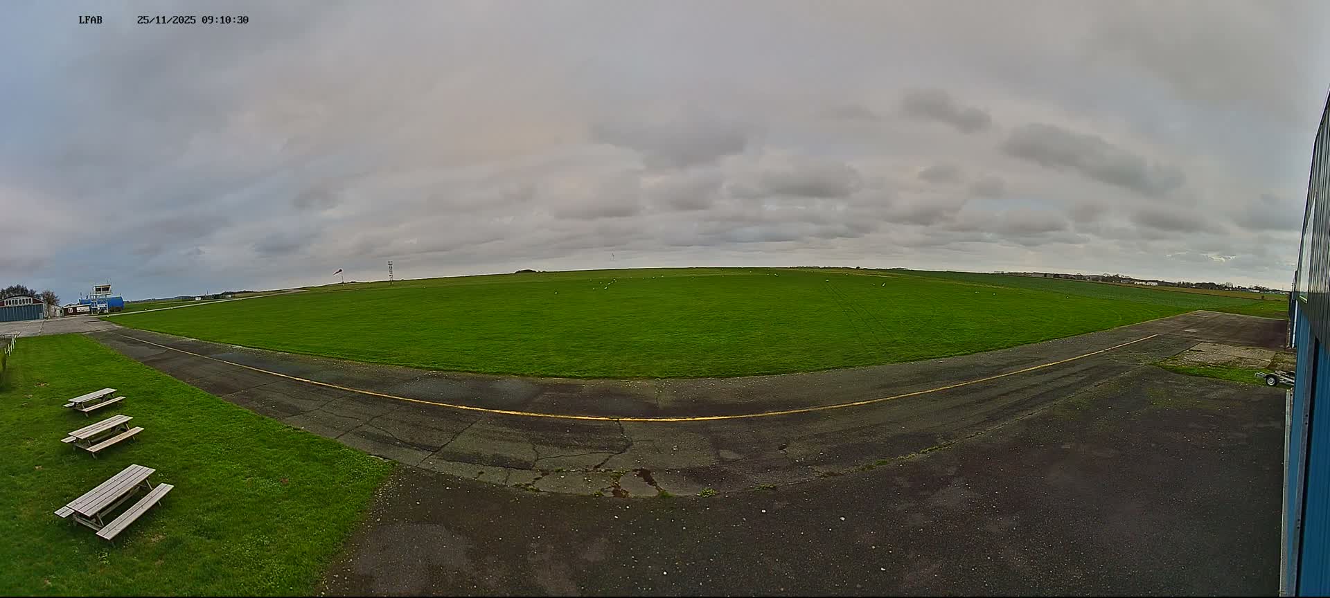 An overcast sky hangs over a panoramic view of a small airfield, featuring a vast green grass field dotted with white objects, a winding paved path, several distant buildings including a control tower, and three picnic tables in the foreground.